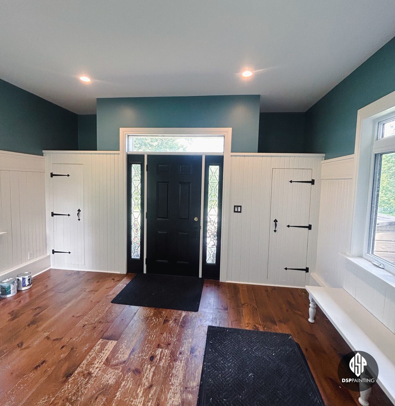 The foyer after painting, teal walls above white beadboard wainscoting around the black front door and two board-and-batten cupboard doors