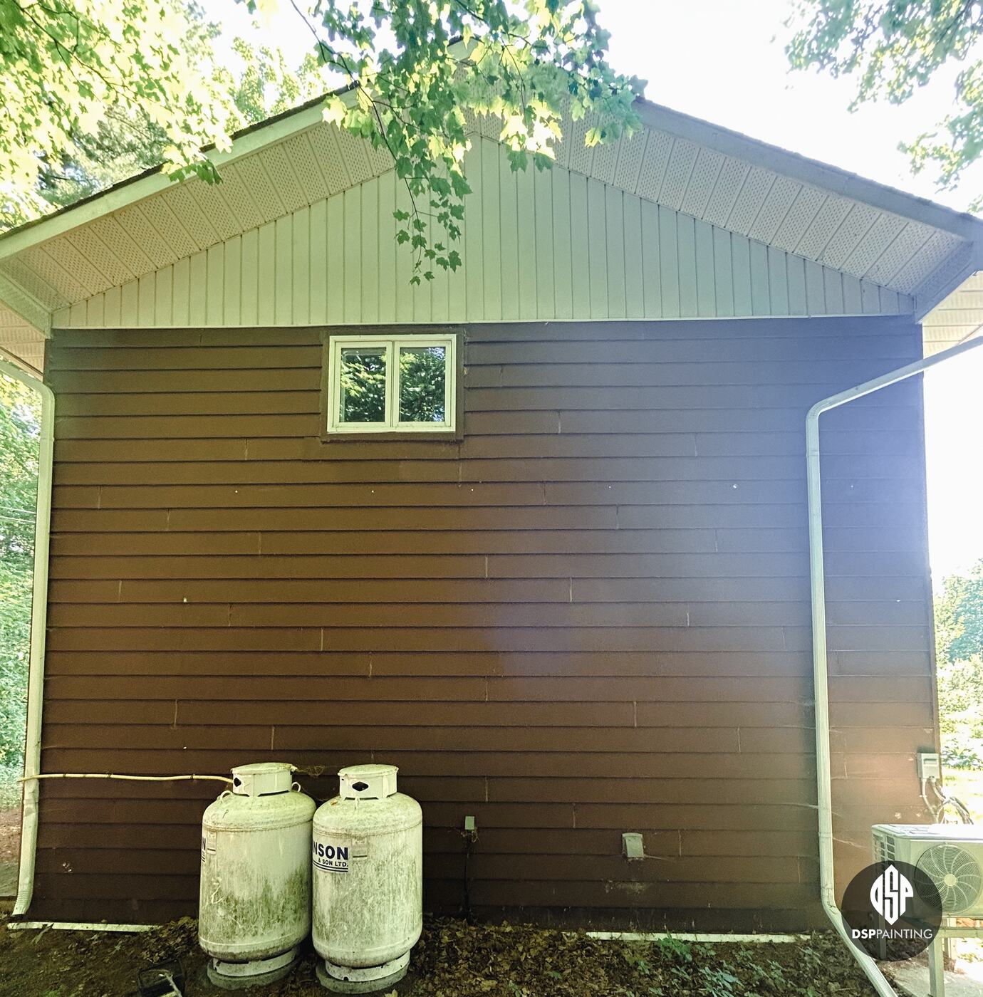 The gable end of the garage, its lap siding red-brown below cream gable siding, with two propane tanks against the wall