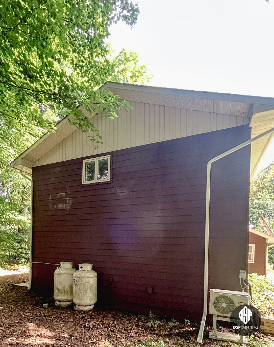 The gable end of the garage in deep red-brown, beside a heat-pump unit and the propane tanks