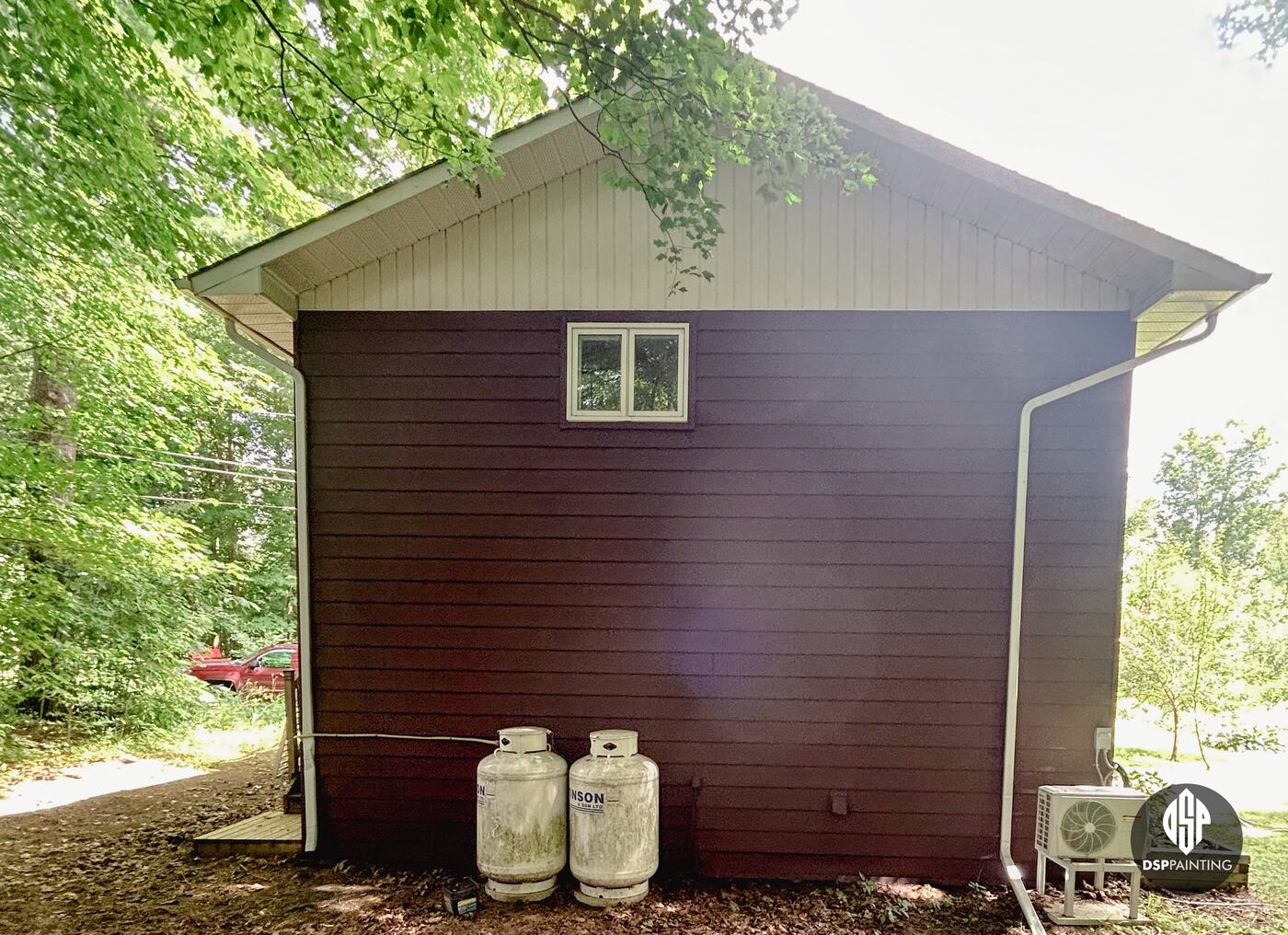 The gable end of the garage, red-brown siding below the cream gable, with the propane tanks and heat pump
