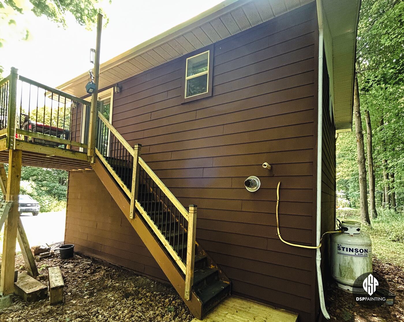 A close view of the back wall, red-brown siding behind the wooden stairs and a propane tank
