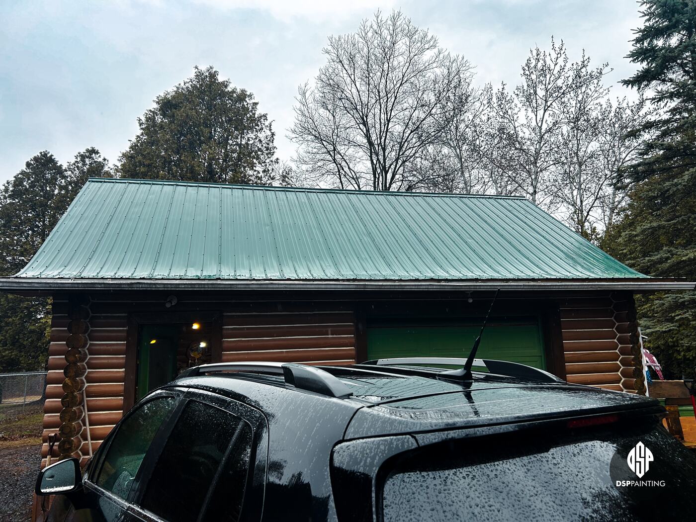 The log garage before the work, its green metal roof faded and dull under a grey sky
