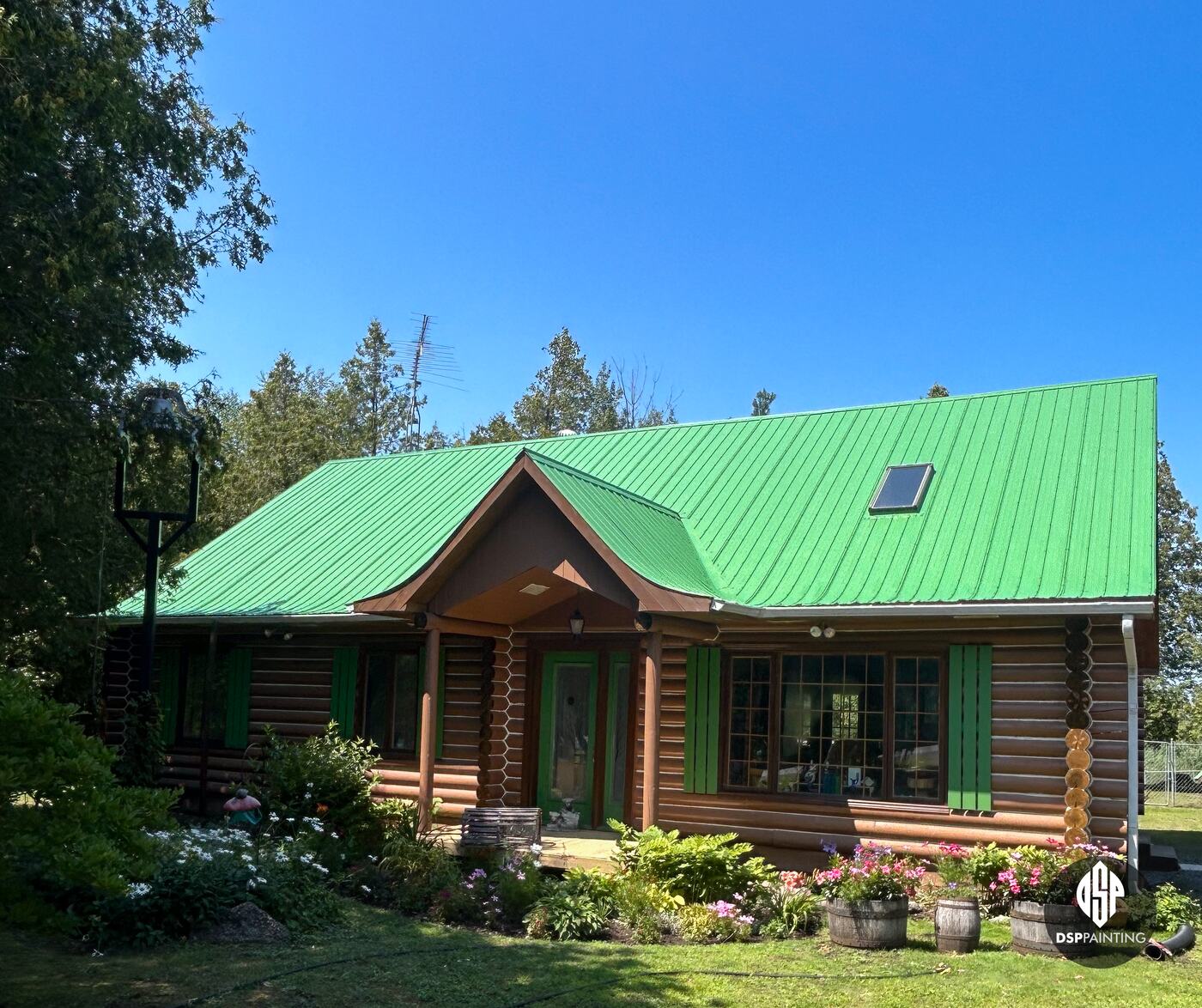 The log house after painting, its metal roof bright green above the log walls, green shutters and flower beds
