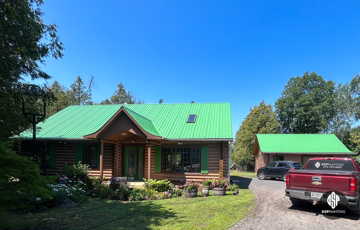 The log house and log garage together, both metal roofs painted bright green, with the DSP Painting truck in the driveway