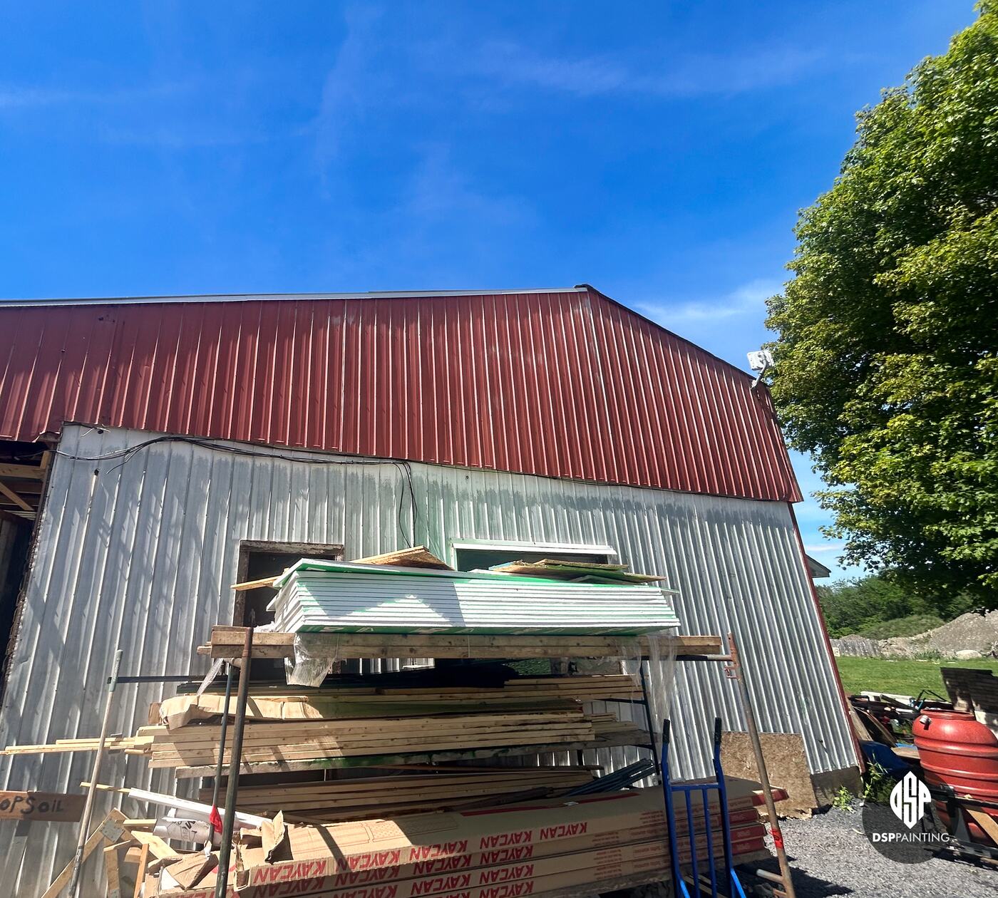 Another side before the work, the red metal roof over white siding, with building materials stacked against the wall