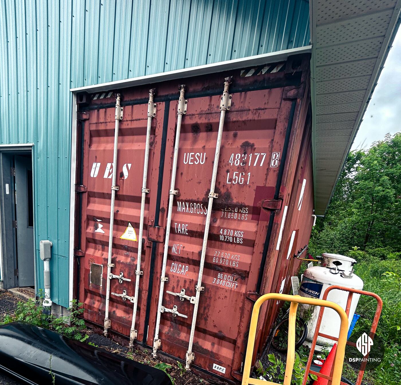 A rusted red shipping container built into the end of a teal steel building, before painting