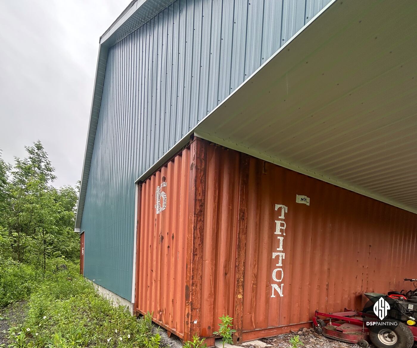 The side of a container under the building’s overhang before painting, rusted red with “Triton” lettering