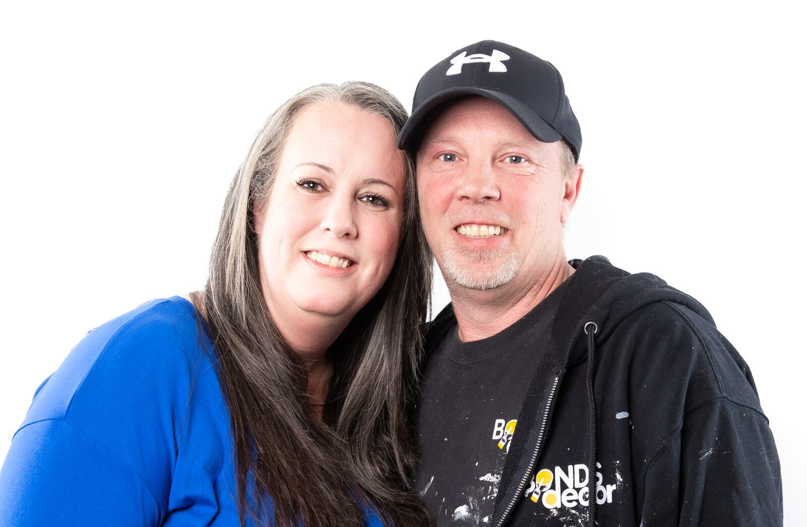 A smiling man in a black cap and paint-spattered work hoodie standing cheek to cheek with a smiling woman in a blue top, against a white studio backdrop.