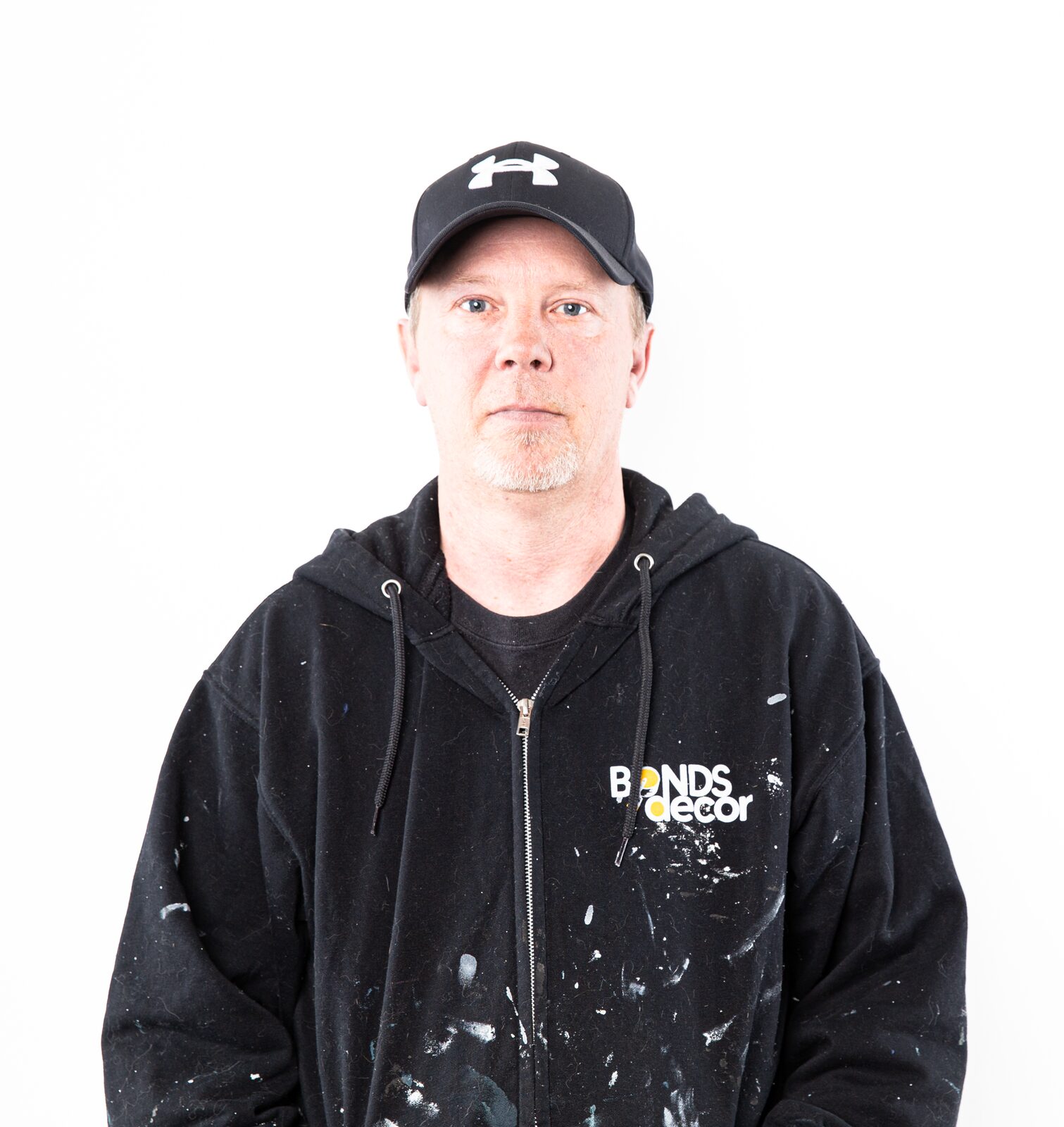 A painter in a black cap and paint-spattered black work hoodie, photographed against a white studio backdrop.